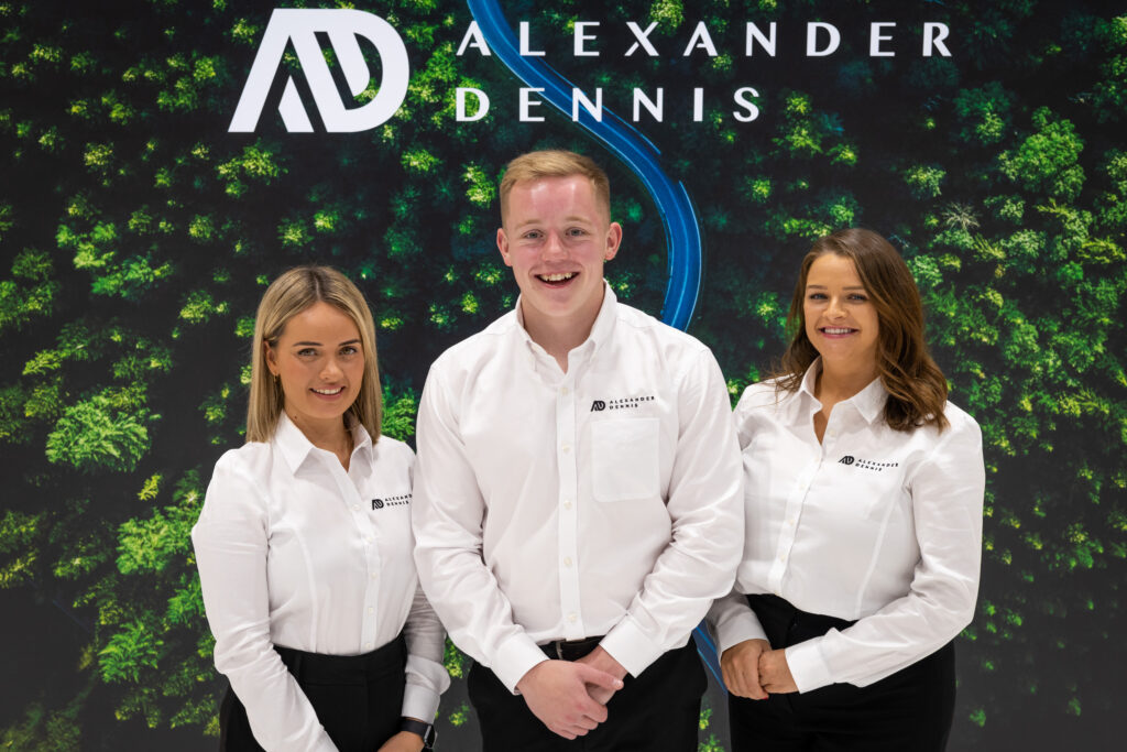 A man and two women stand in front of an Alexander Dennis logo