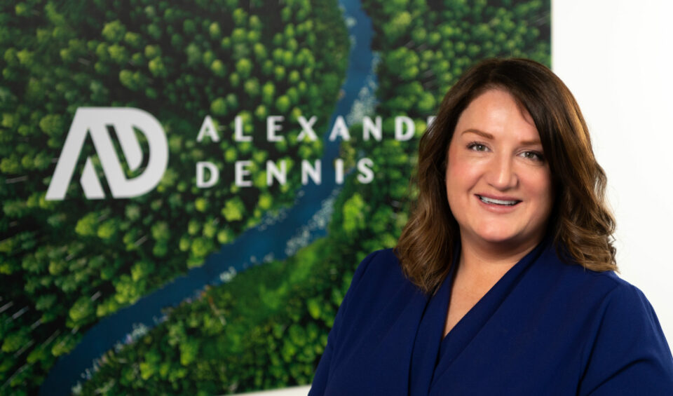 Portrait photo of a white woman in front of a picture that shows a birds-eye view of a forest and river with the Alexander Dennis logo overlaid.