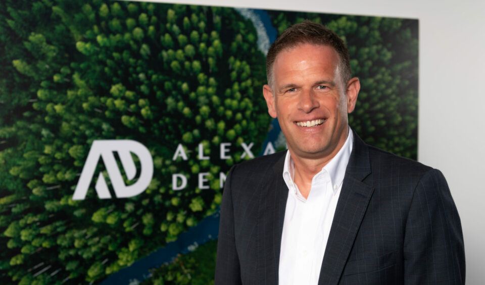 Portrait photo of a white man in front of a picture that shows a birds-eye view of a forest and river with the Alexander Dennis logo overlaid.