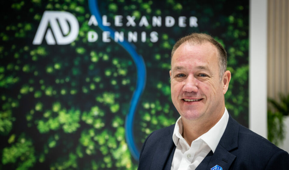 Portrait photo of a middle-aged white man in front of a picture that shows a birds-eye view of a forest and river with the Alexander Dennis logo overlaid.