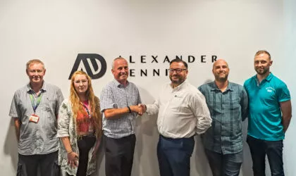 Six white people stand in front of a white wall with the Alexander Dennis logo on it. The two men in the centre shake hands.