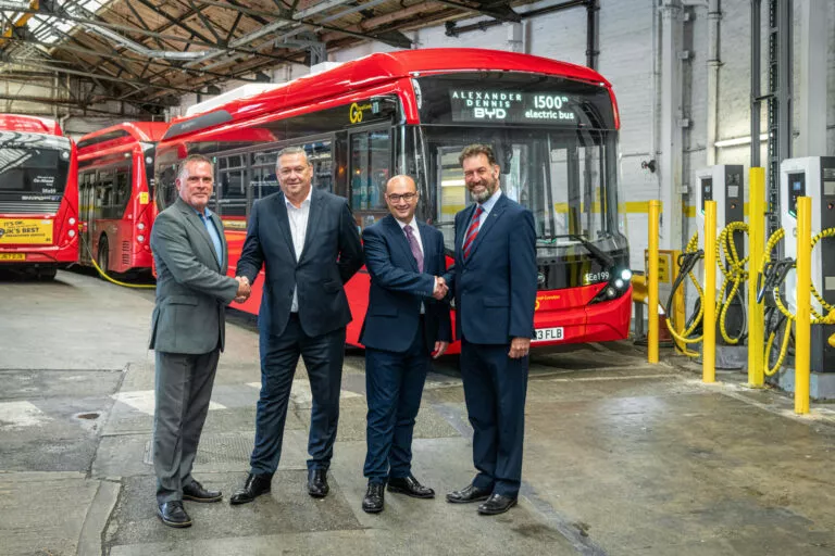 Four men in suits shake hands in front of a red single deck bus which has 'Alexander Dennis BYD 1500th electric bus' on its display