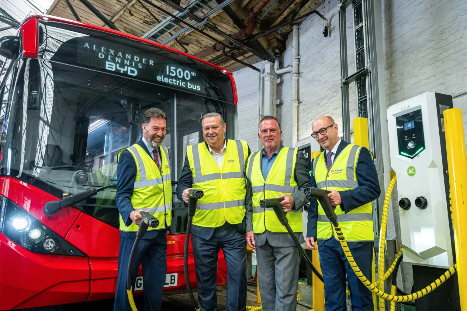 Four men in high visibility jackets hold electric vehicle charging plugs in front of a red single deck bus which has 'Alexander Dennis BYD 1500th electric bus' on its display