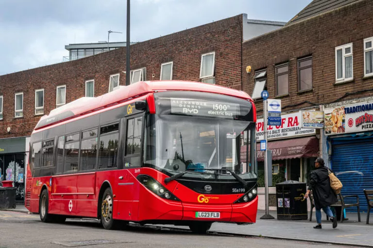 A red single deck bus which has 'Alexander Dennis BYD 1500th electric bus' on its display stands at a bus stop in front of a row of shops