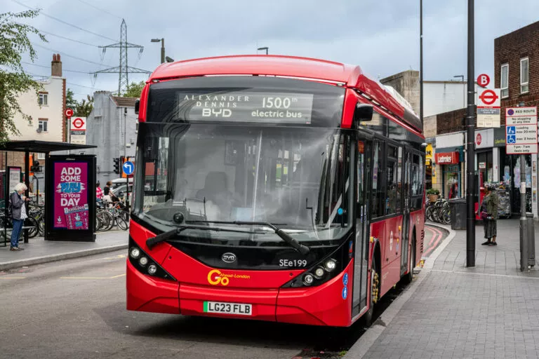 A red single deck bus which has 'Alexander Dennis BYD 1500th electric bus' on its display stands at a bus stop